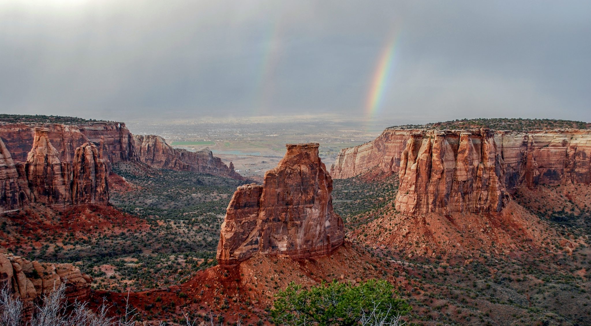 landscape photo of Colorado National monument, red rock expanses, deep canyons, and sandstone towers with 2 diagonal rainbows in the distance
