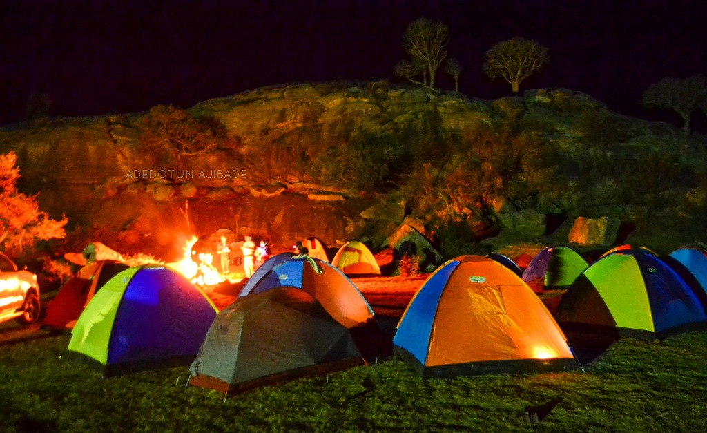 nighttime. red rock expanses with multiple colorful tents in the foreground. People are standing around a fire pit.