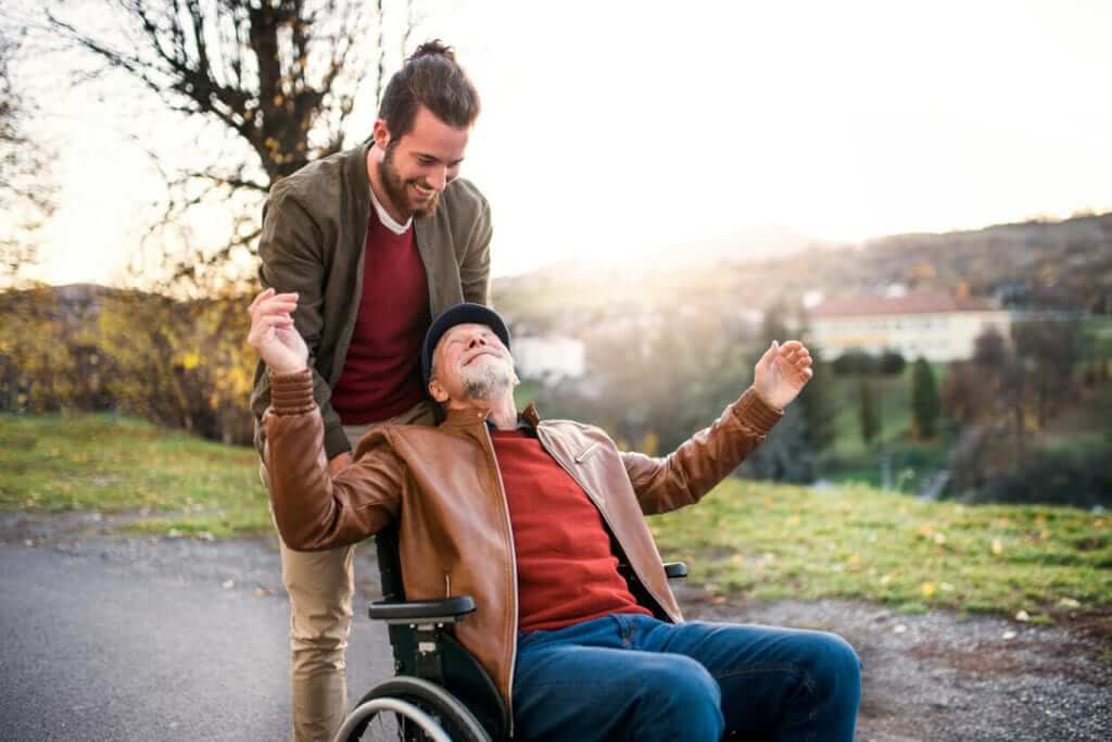 caucasian adult senior male seated wearing blue jeans, a red sweater, tan jacket and a black hat, leaning back in his wheelchair with his arms spread, looking up at a younger adult male standing behind him, wearing tan pants, a red shirt and a tan jacket looking down at the seated man outdoors on a paved path with grass, trees, a body of water, and the sunset in the background