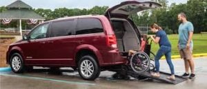 red minivan with rear wheelchair ramp. caucasian adult couple is pushing male minor into the van in his wheelchair