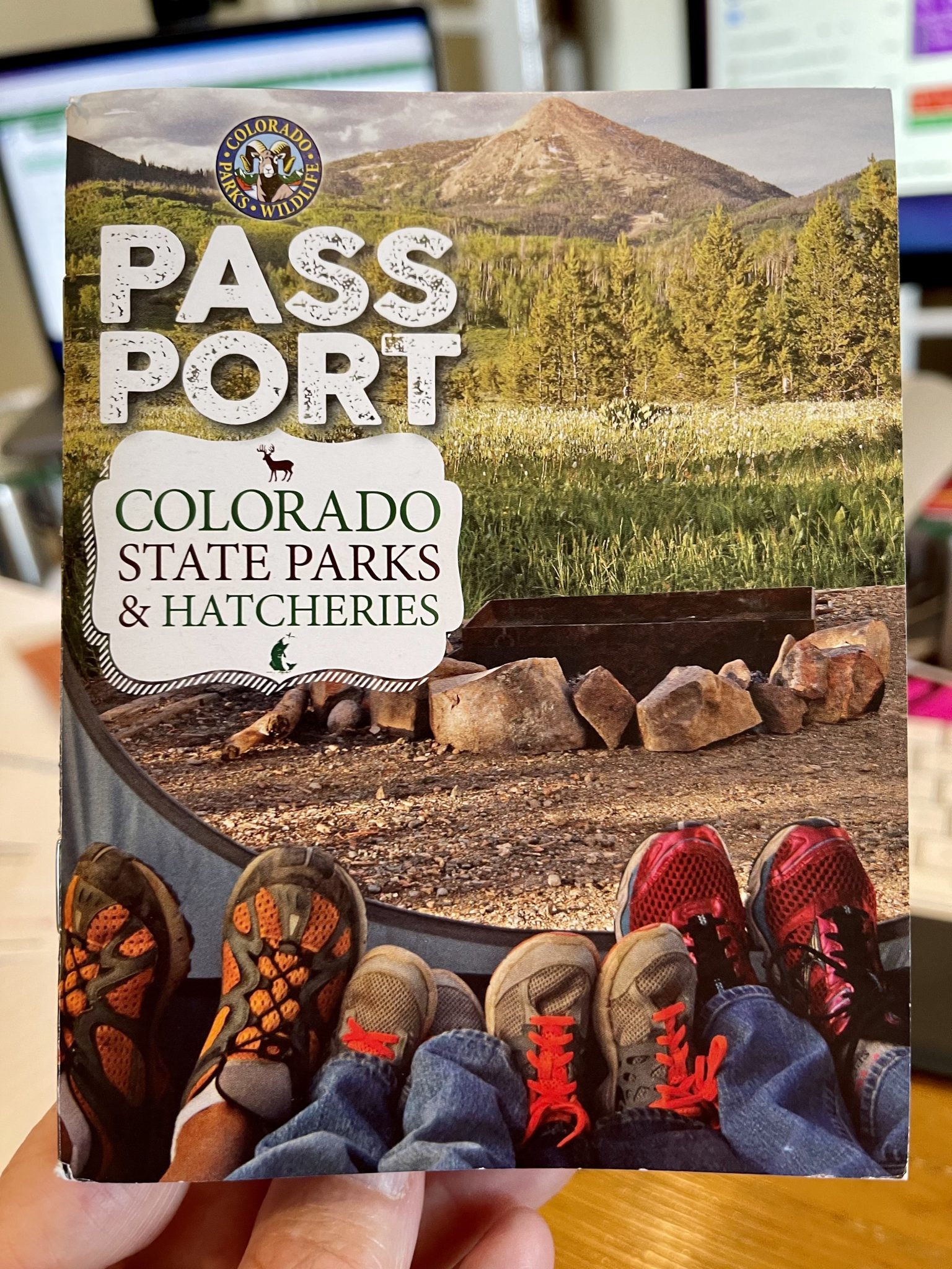 Colorado Parks and Wildlife Passport cover. photo of people's legs at the interior edge of a tent looking out toward a fire ring, a meadow, trees and a mountain in the distance. [Text: Colorado Parks and Wildlife Passport. Colorado state parks & hatcheries