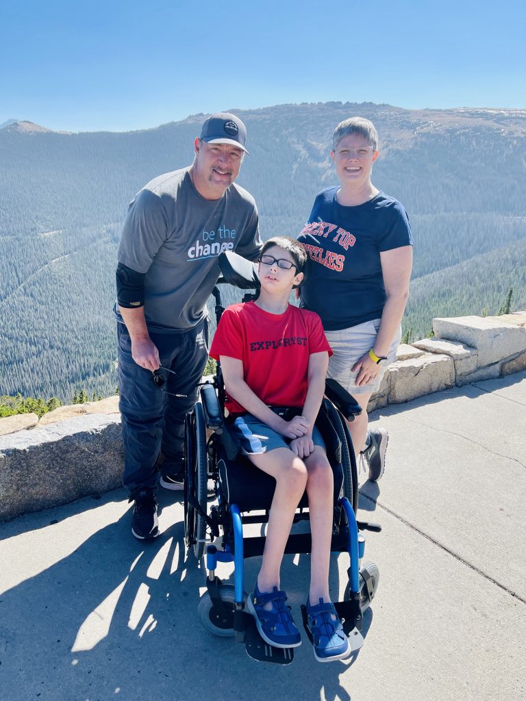 adult male and adult female standing behind a male youth in a wheelchair. The background is a mountainous expanse in Rocky Mountain National Park