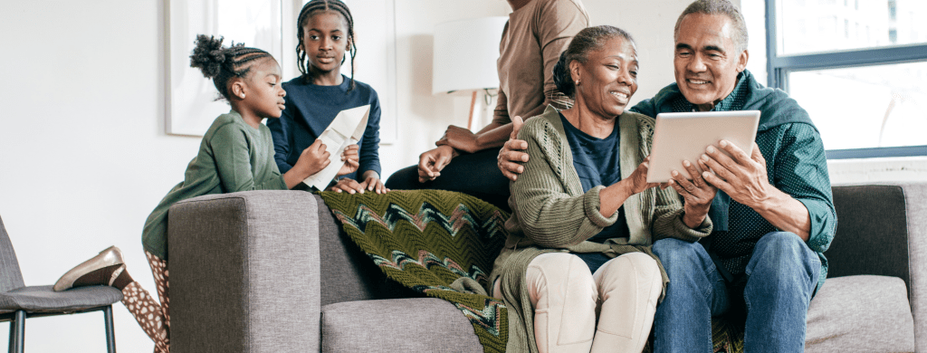 older Black couple looking at an ipad on a couch, young Black youths behind the couch looking at an ipad