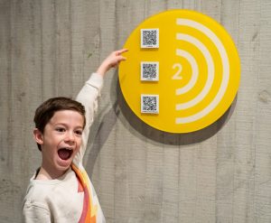 child next to a yellow sign with 3 QR codes